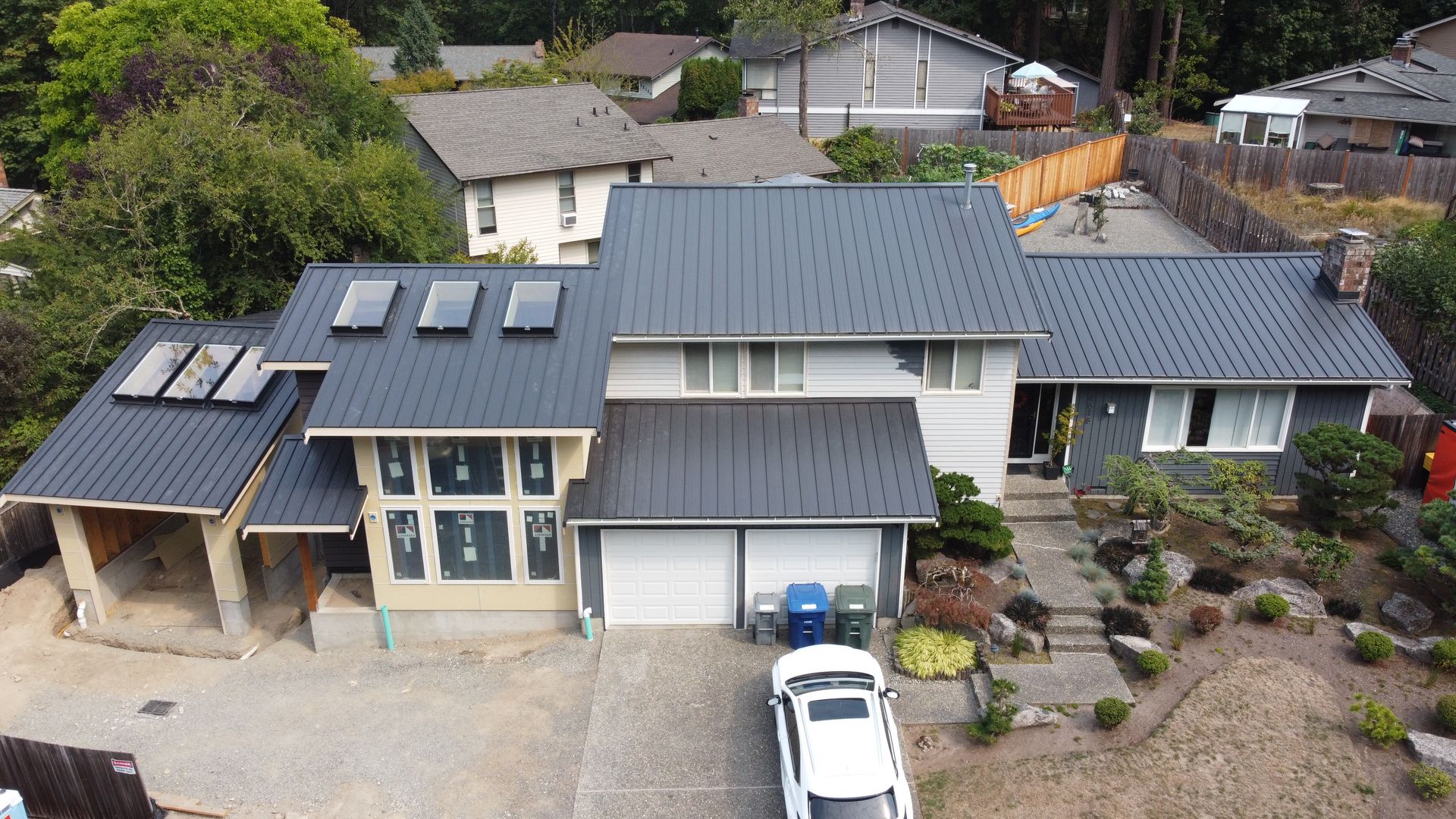 Aerial view of a modern gray-roofed house with skylights, front driveway, and landscaped yard.