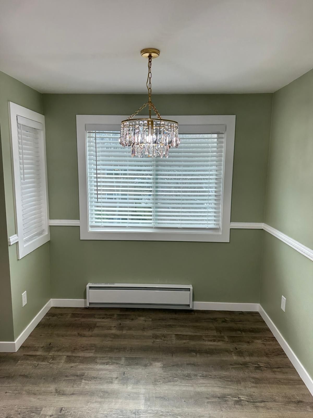 Empty dining room with sage green walls, white trim, chandelier, and window blinds on wood-look floor.