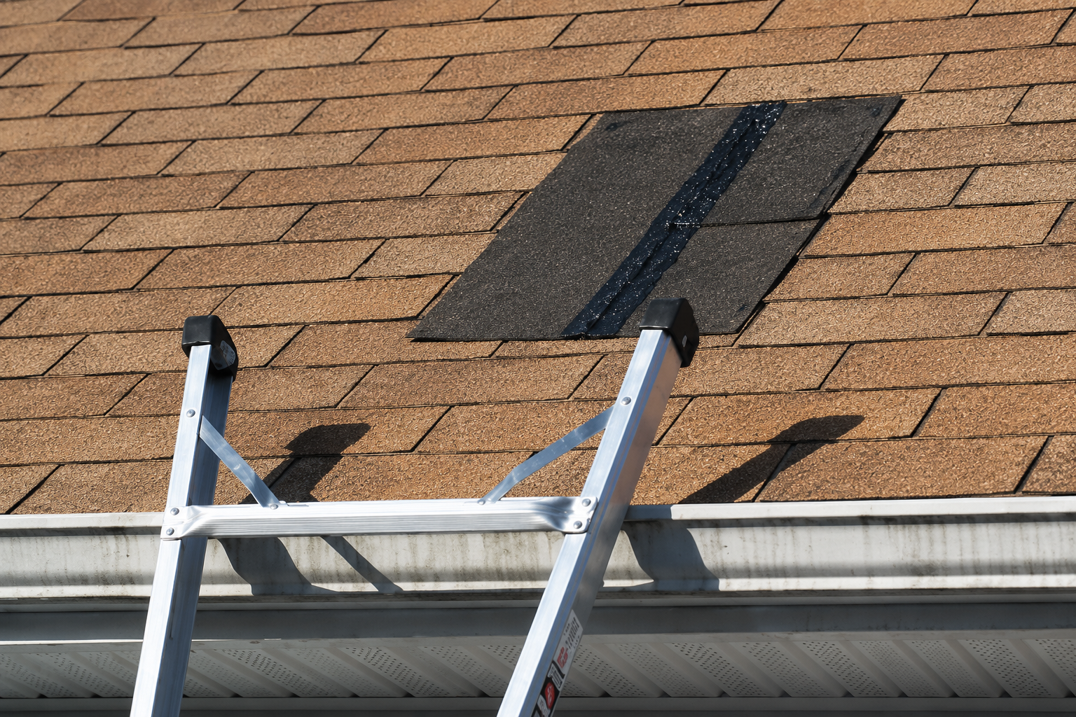 Roof ladder leaning against shingled roof near a dark rectangular patch repair
