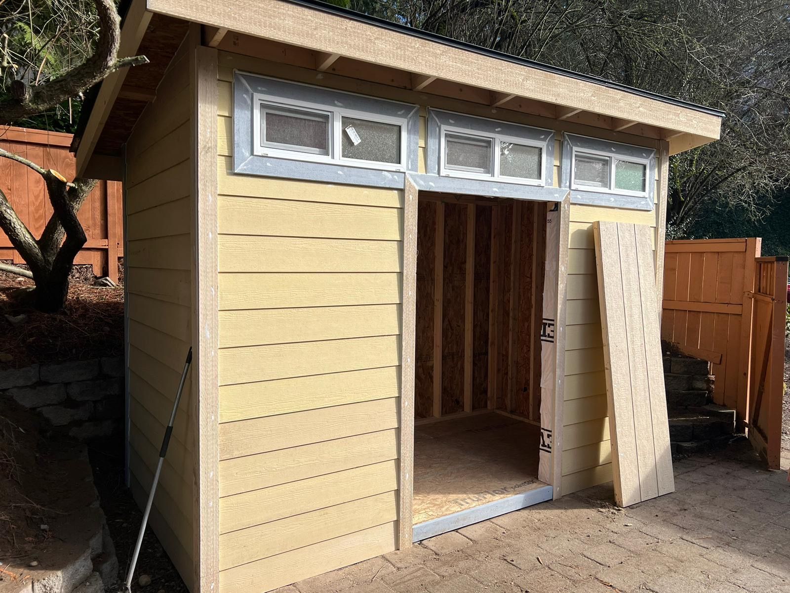 Small yellow wooden shed with open double doors and windows along the top, set outdoors on a patio.