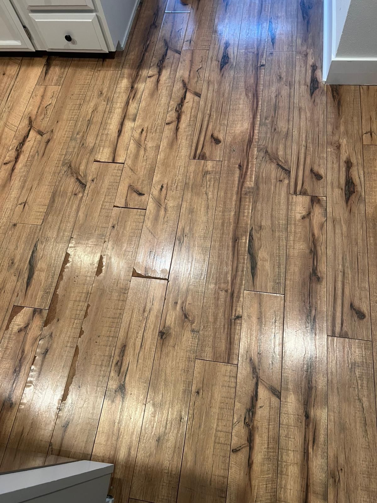 Wood-look laminate flooring in a hallway or kitchen area with light reflections and white cabinetry edges.