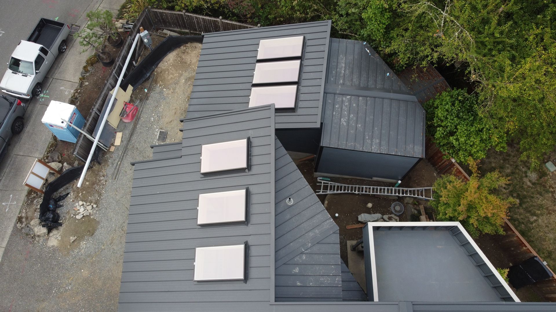 Aerial view of a gray-roofed building with skylights beside trees and a gravel area.