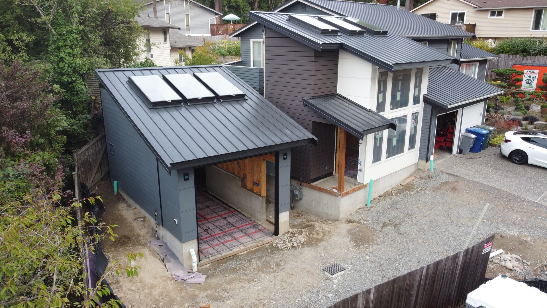 Modern house with dark roof and solar panels beside a gravel driveway in a wooded neighborhood