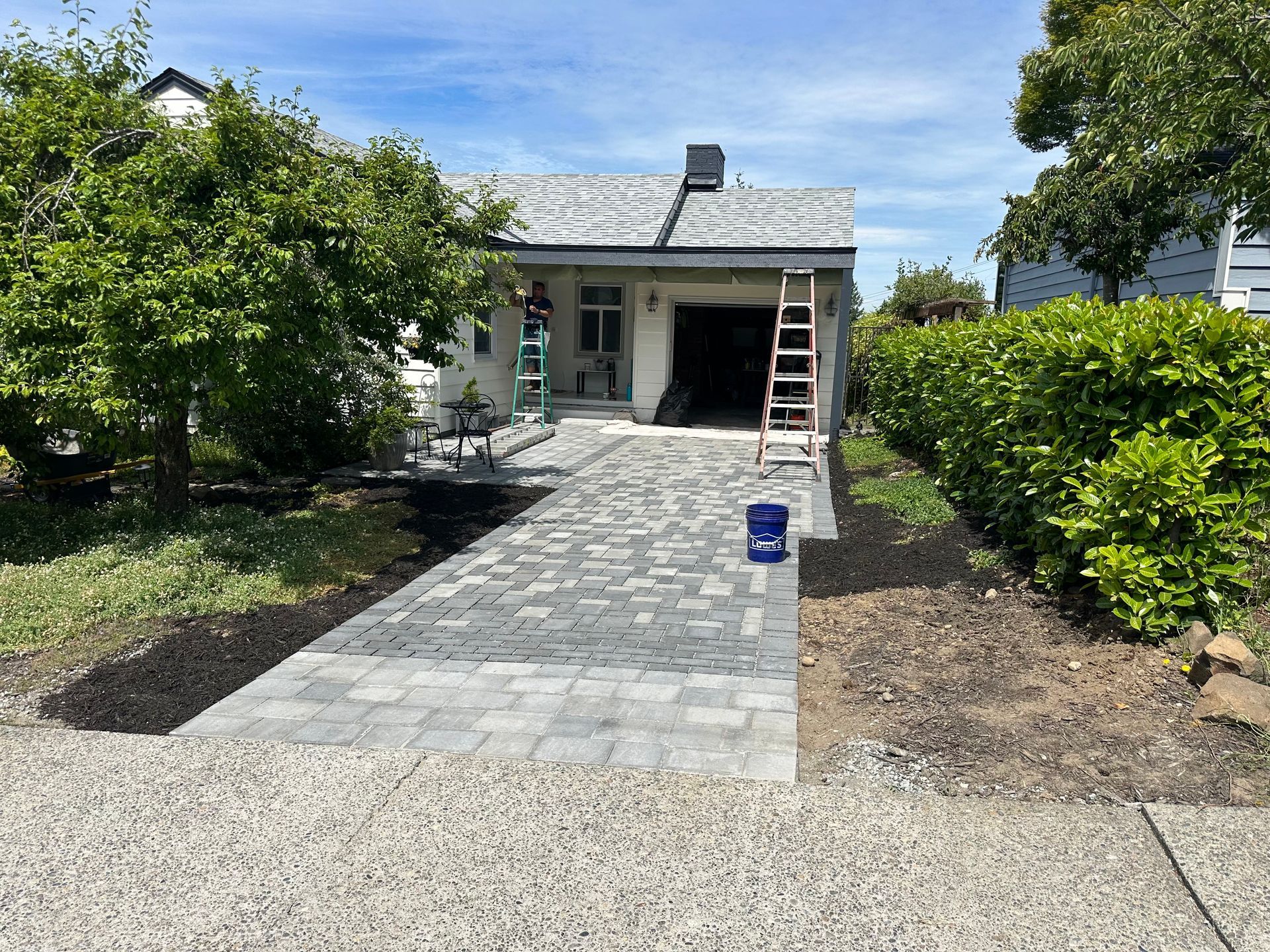 Front walkway to a house under construction, with a ladder on the porch and a bucket on the paver path.