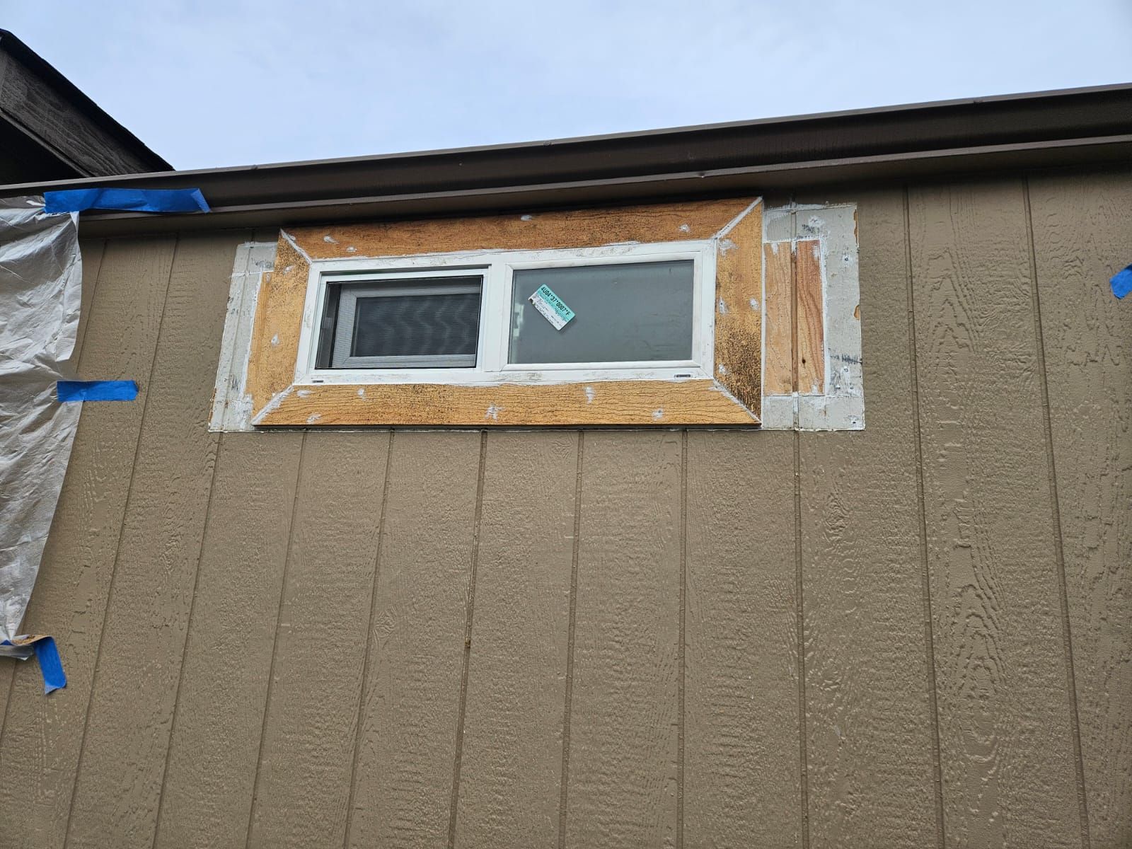 Small window set into tan exterior siding with unfinished trim and blue tape around it