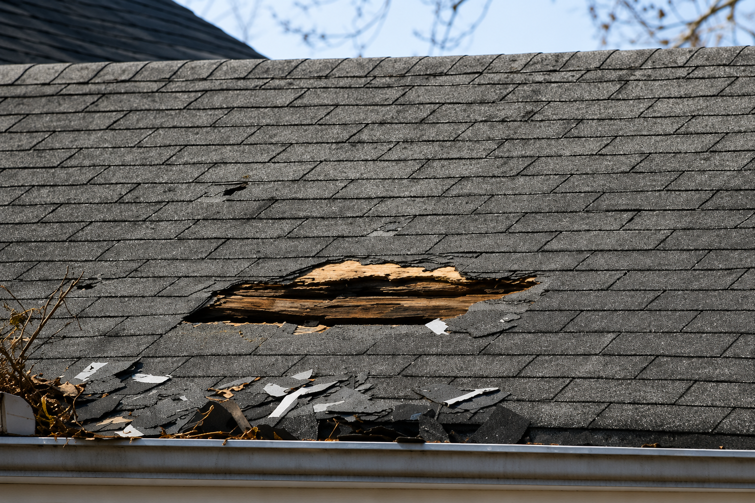 Damaged shingle roof with a large hole exposing the wooden underlayment and debris on the edge