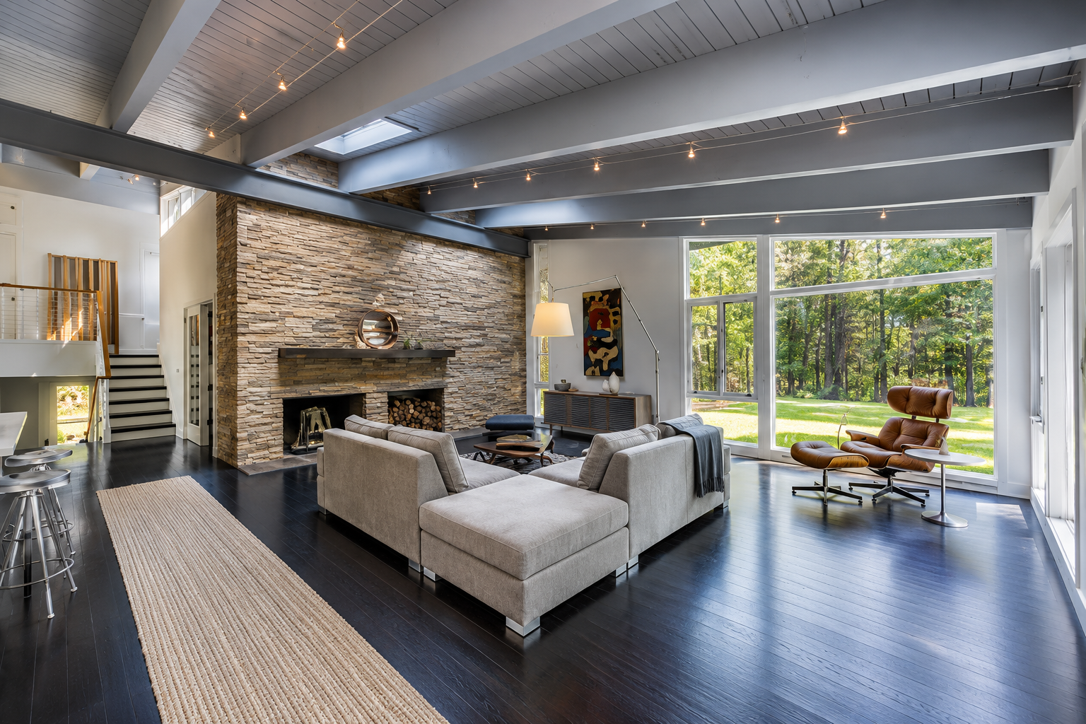 Modern living room with stone fireplace, gray sectional, dark wood floor, and large windows to a green backyard