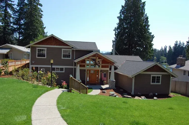 Suburban house with brown siding, green lawn, curved walkway, and tall evergreen trees under a clear sky