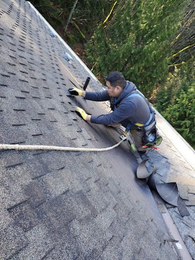Worker on a steep shingled roof using a harness and tools to repair a section near the ridge.