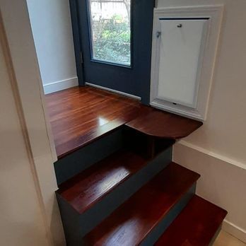 Narrow dark-stained staircase leading to a small landing beside a white cabinet and window in a bright room