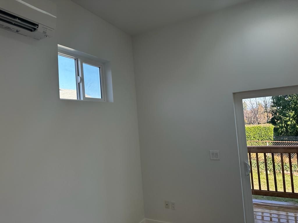 Empty white room with a small window, doorway, and view of a balcony and greenery outside