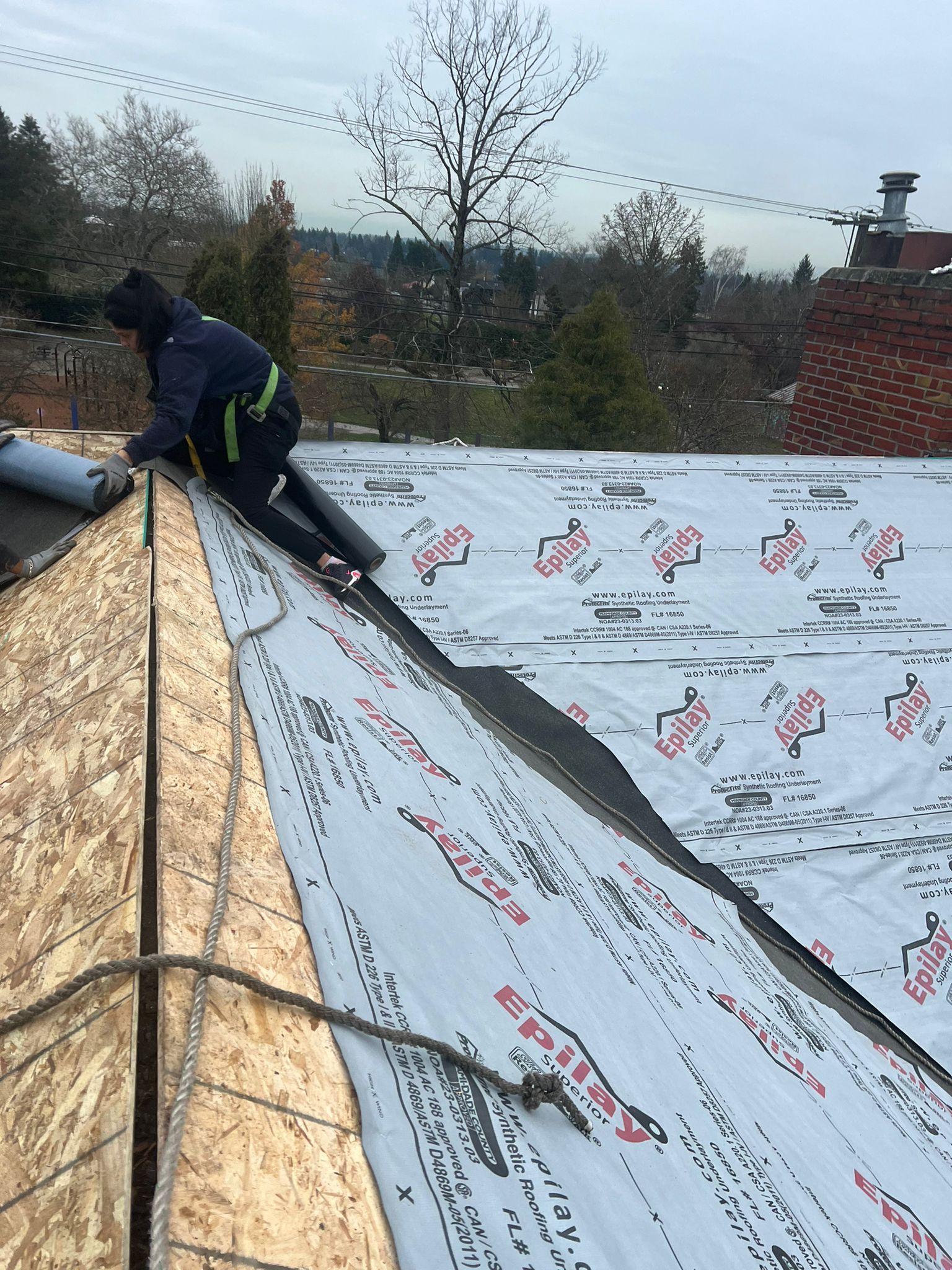 Worker installing roofing underlayment on a sloped roof, with overcast sky and trees in the background