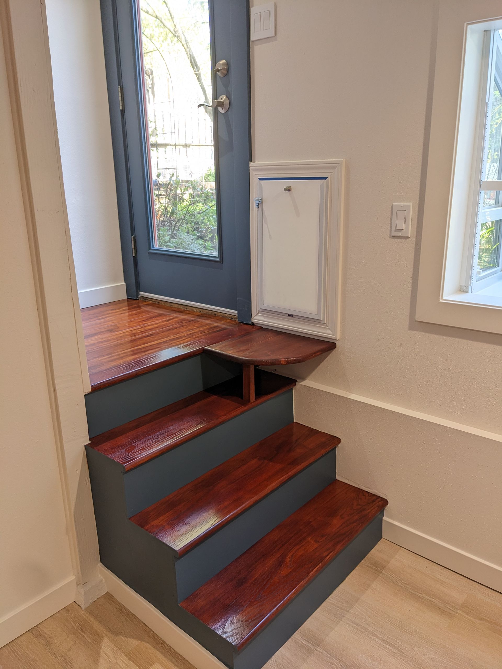 Indoor stairs with dark red carpet and blue trim leading to a gray front door in a bright entryway