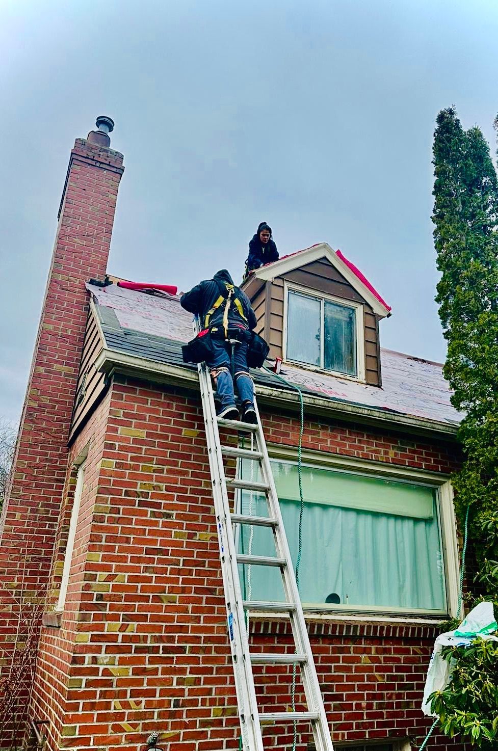 Worker on a ladder repairing the roof of a brick house with green-trimmed windows.