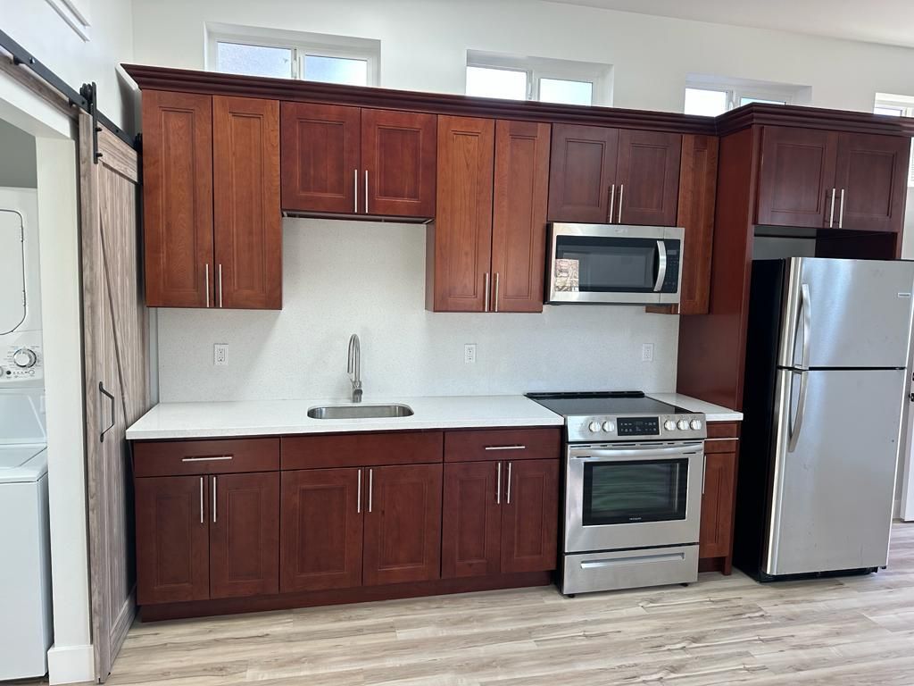 Compact kitchen with dark wood cabinets, white countertops, stainless steel appliances, and a window above the sink