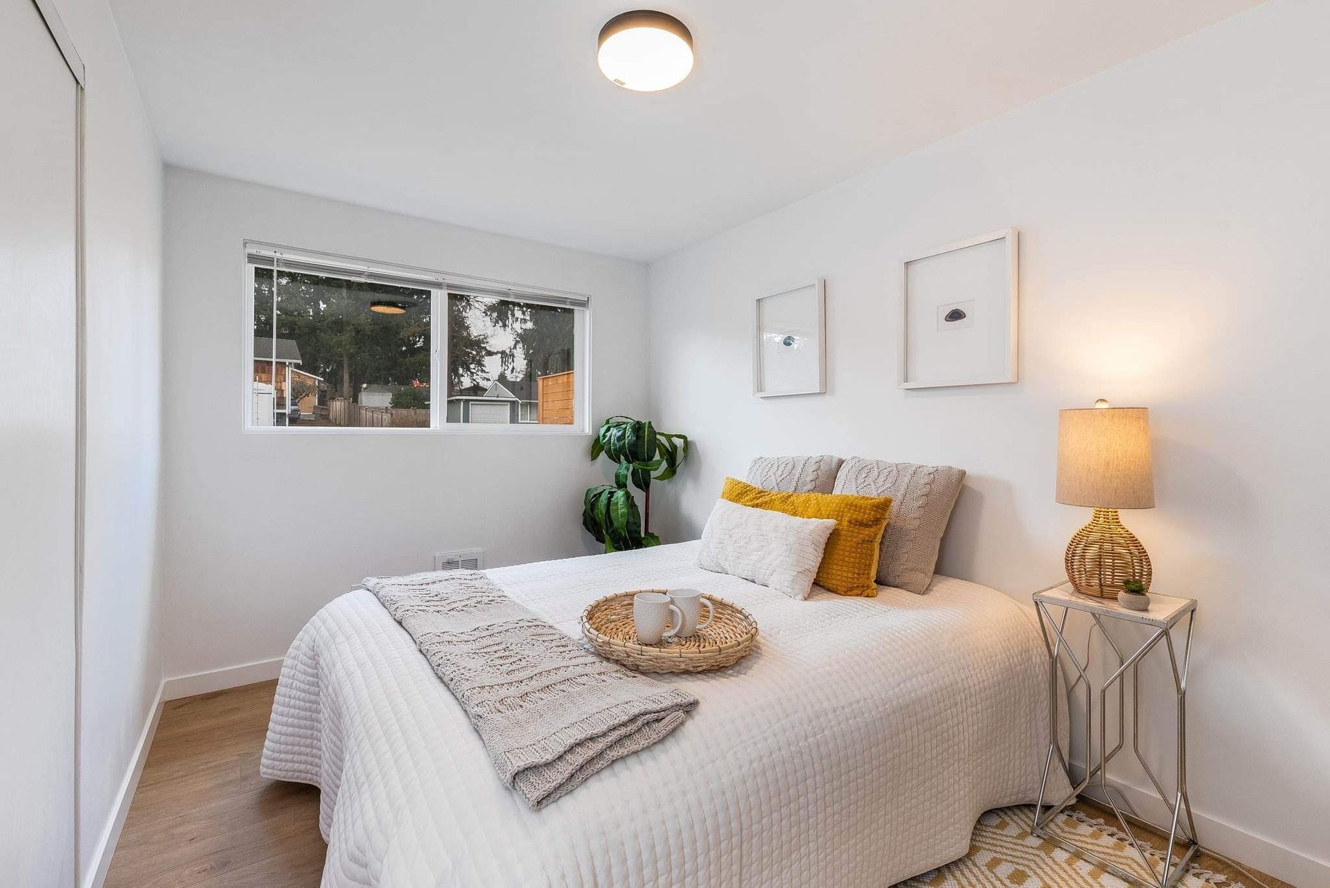 Bright bedroom with white bedspread, wooden floor, window, plant, and bedside lamp