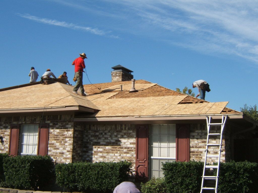 Roofers working on a shingle roof beside a brick house, with a ladder on the right side.