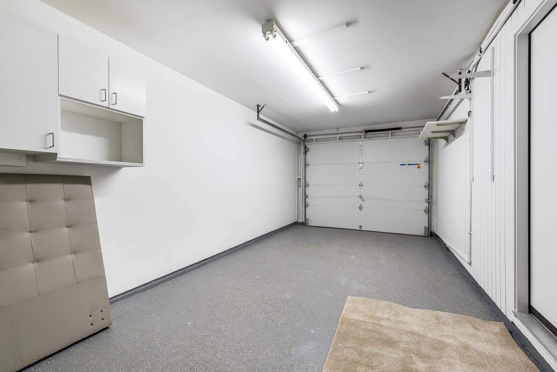 Empty garage with white walls, gray speckled floor, overhead lights, and a closed white garage door