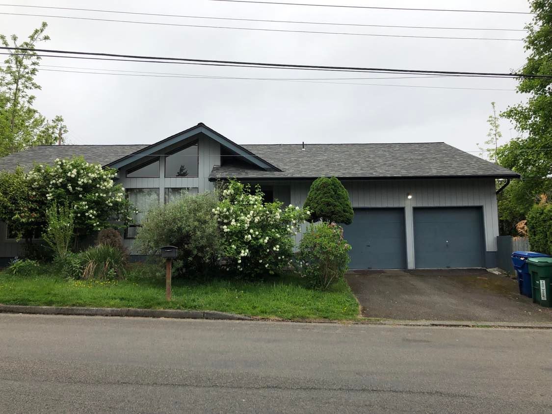 Gray house with two garages, overgrown shrubs, and a driveway on a suburban street