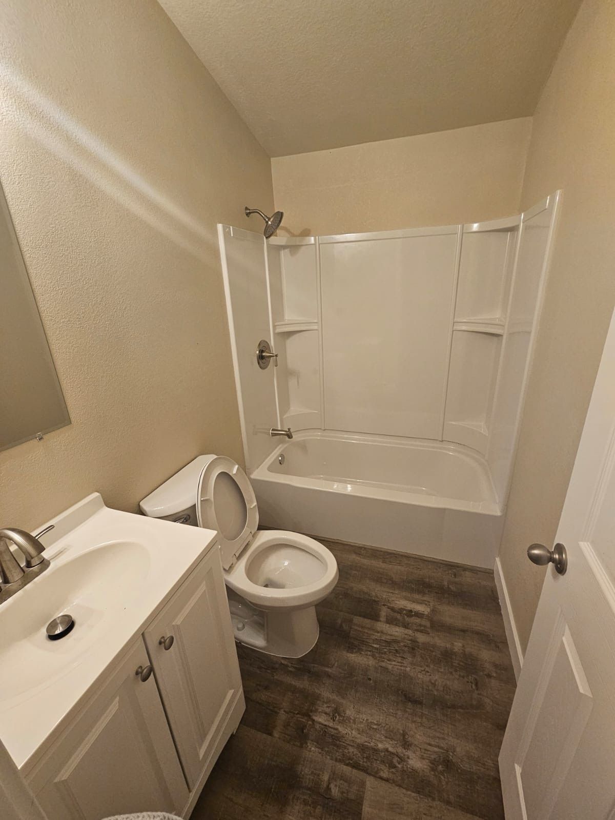 Small beige bathroom with white vanity, toilet, and shower-tub with sliding glass doors and wood-look floor.