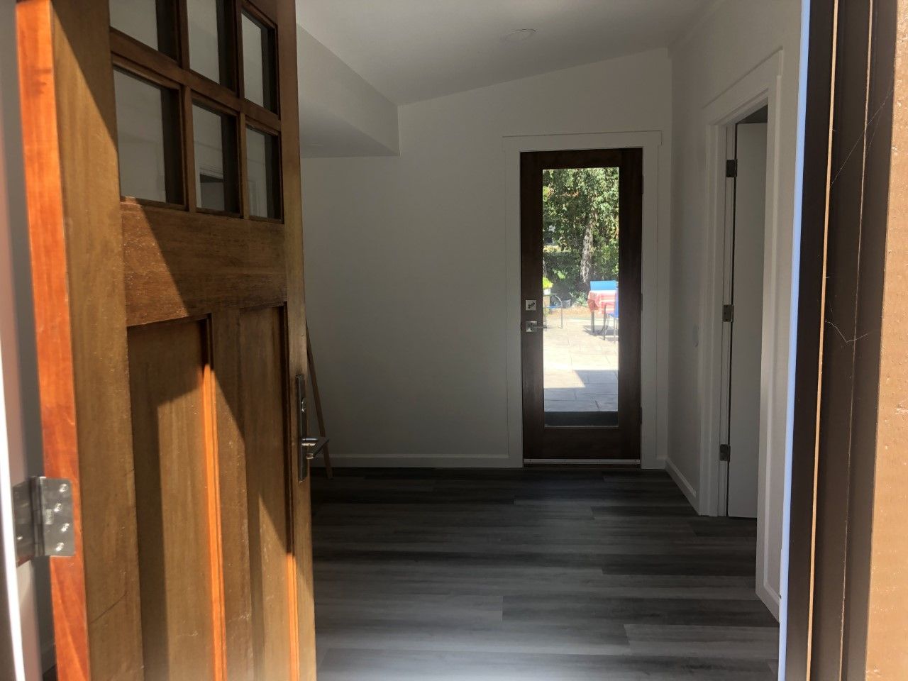 Empty hallway with dark wood floor, white walls, and an open wooden door by a bright entryway.