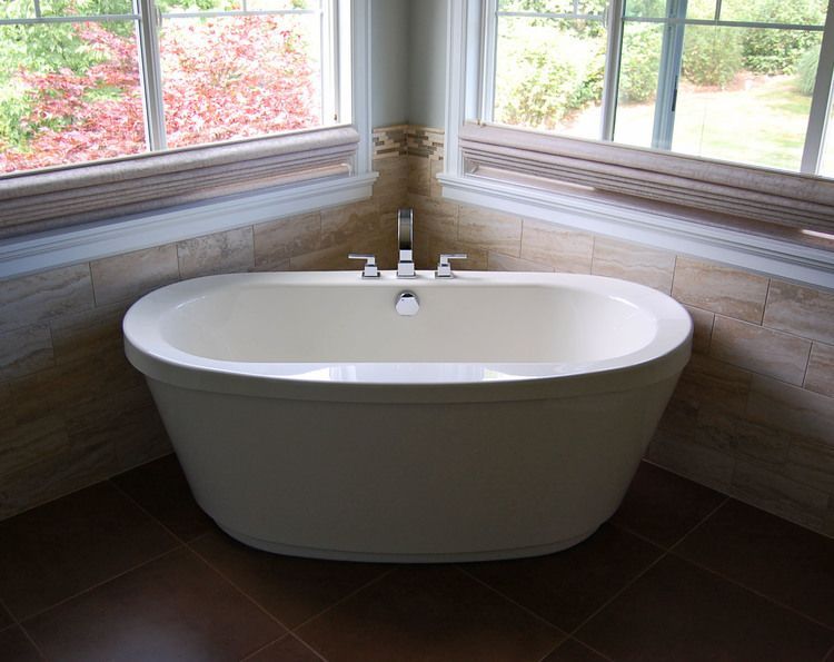 Freestanding white bathtub by corner windows in a bright bathroom with outdoor greenery visible