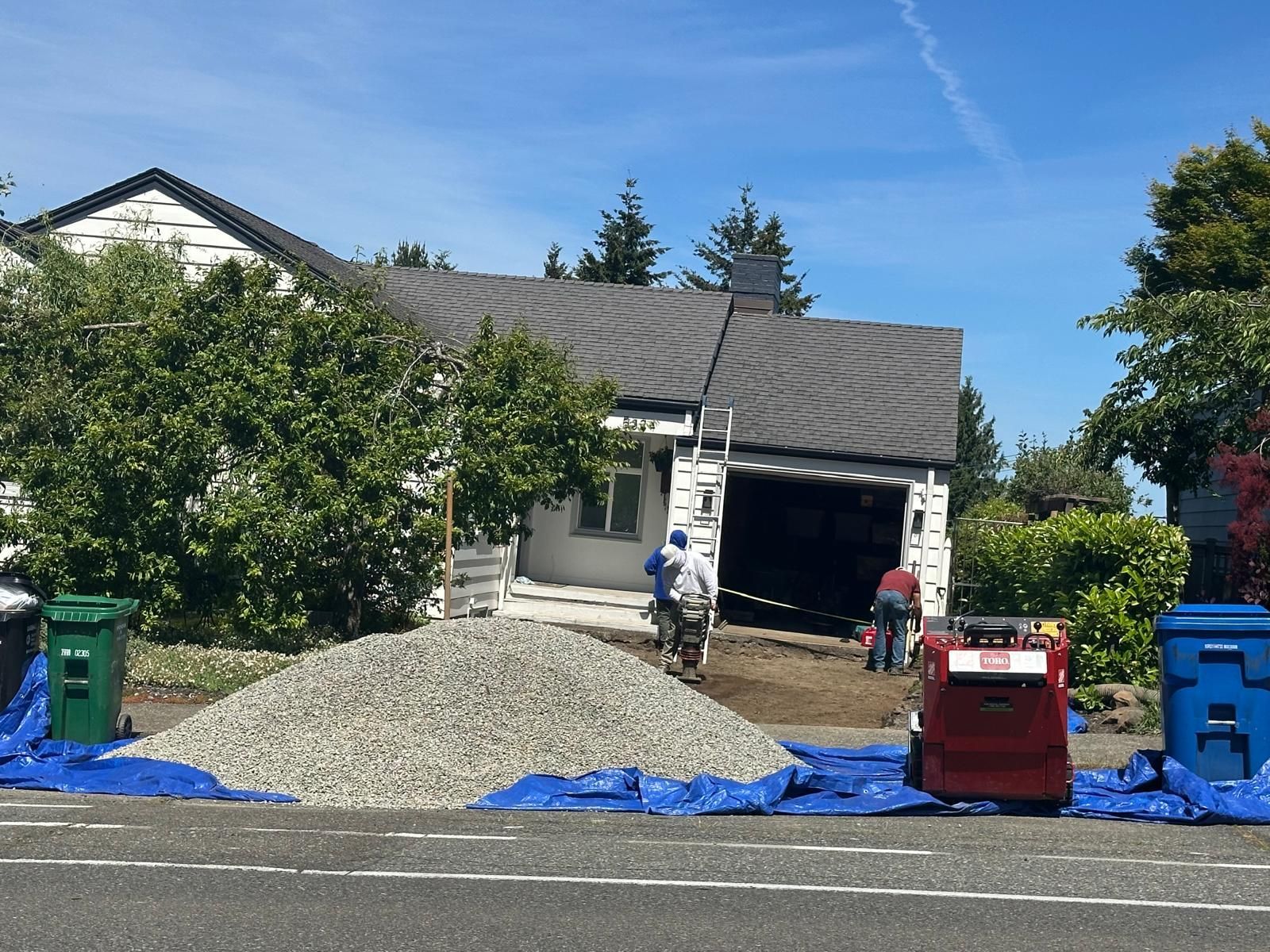 Workers with a gravel pile and blue tarp in front of a house garage, with red and blue bins nearby