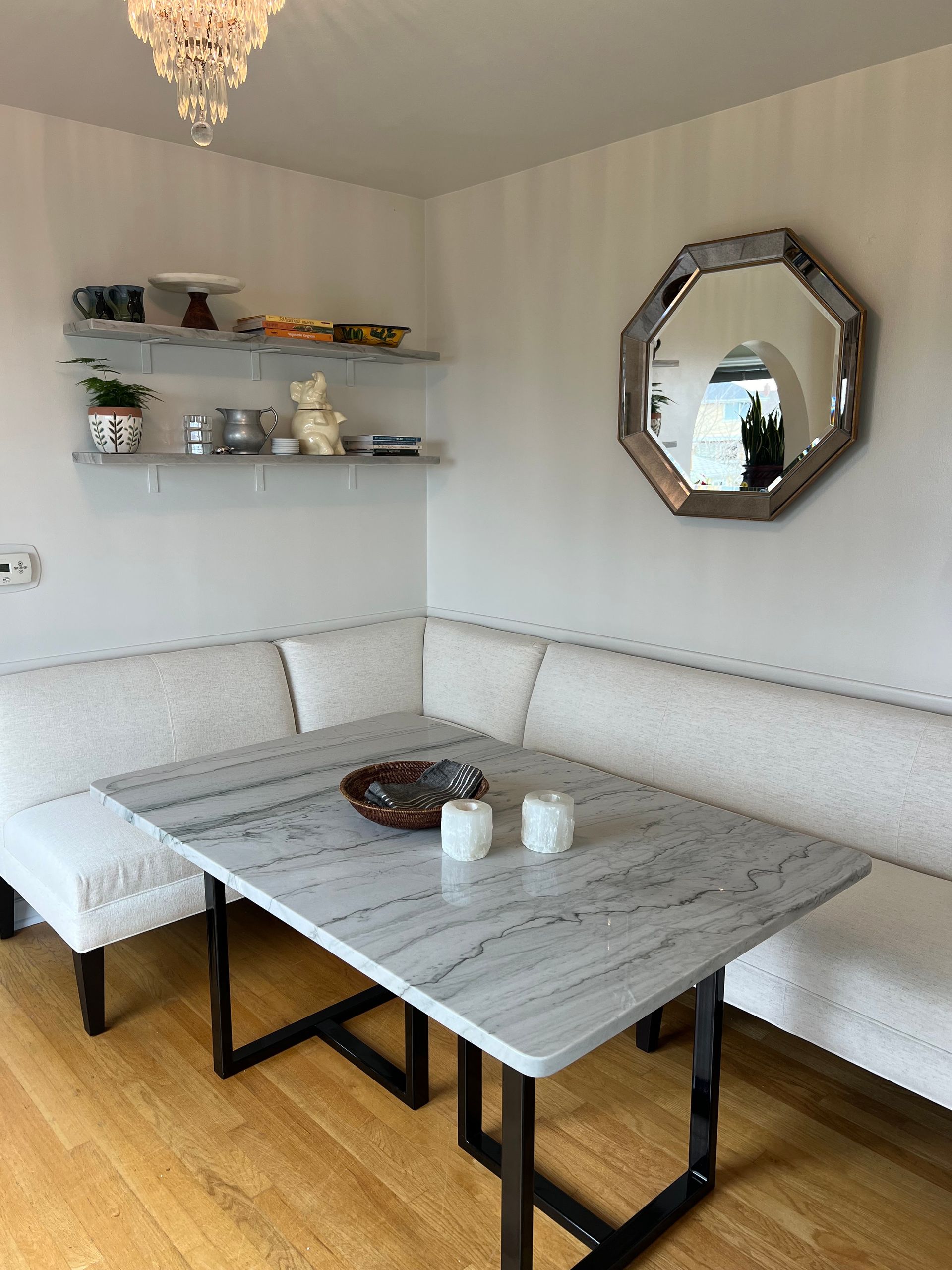 Cozy dining nook with white sectional bench, gray table, wall shelves, and octagonal mirror