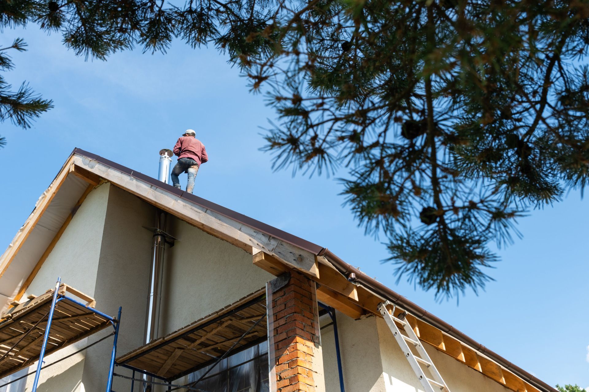 Worker on a roof beside a house under a blue sky, with tree branches in the foreground