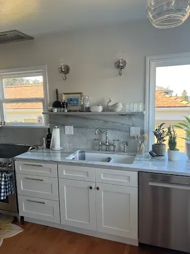 Bright kitchen with white cabinets, marble countertop, sink, stainless dishwasher, and two windows overlooking rooftops
