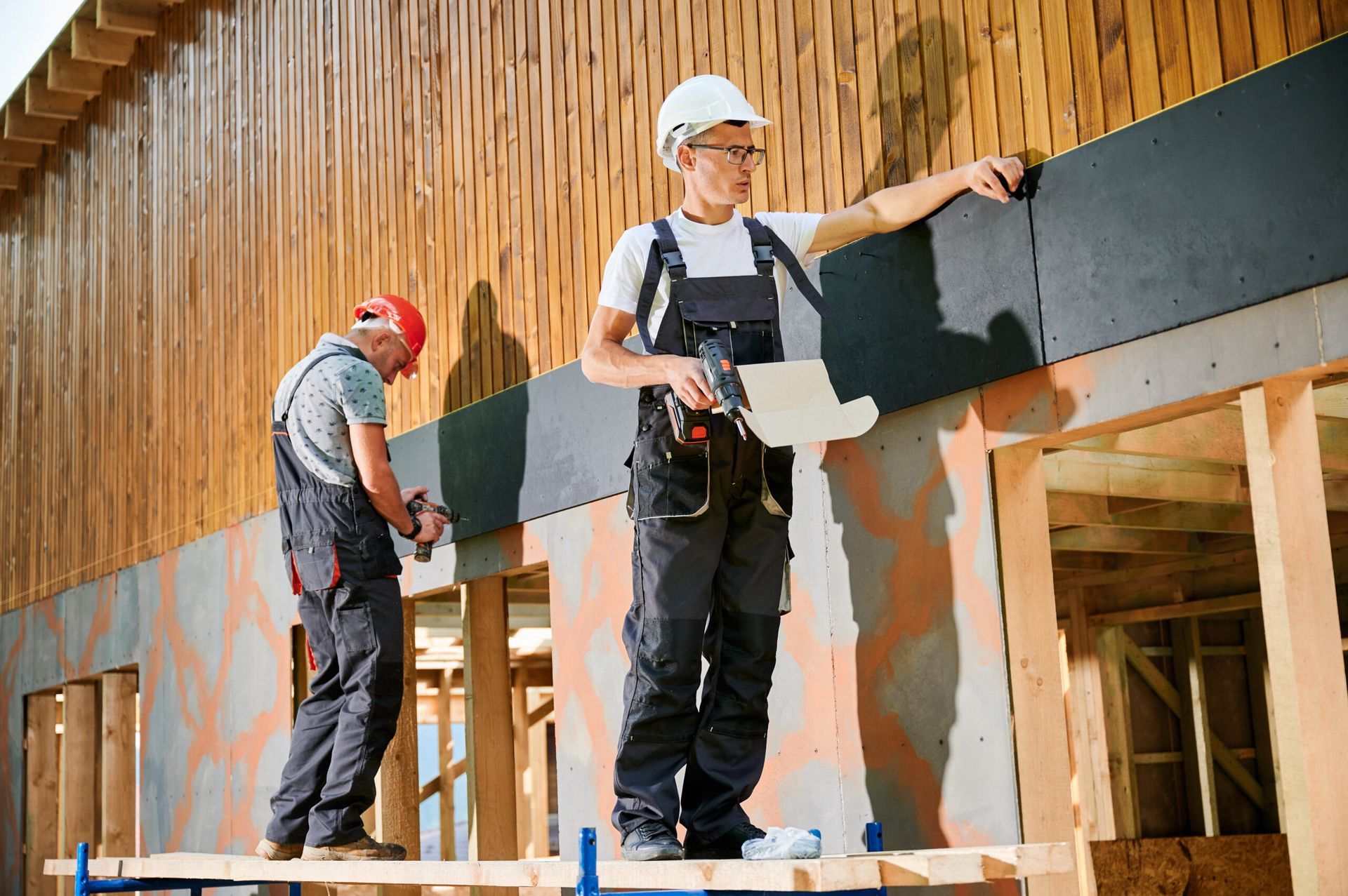 Two construction workers installing panels on a wooden building frame outdoors.