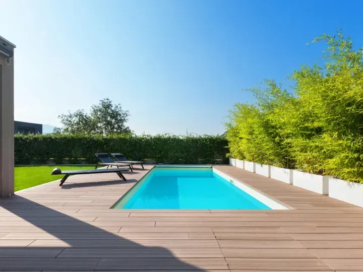 Modern backyard with turquoise swimming pool, wooden deck, lounge chairs, and green hedges under a blue sky