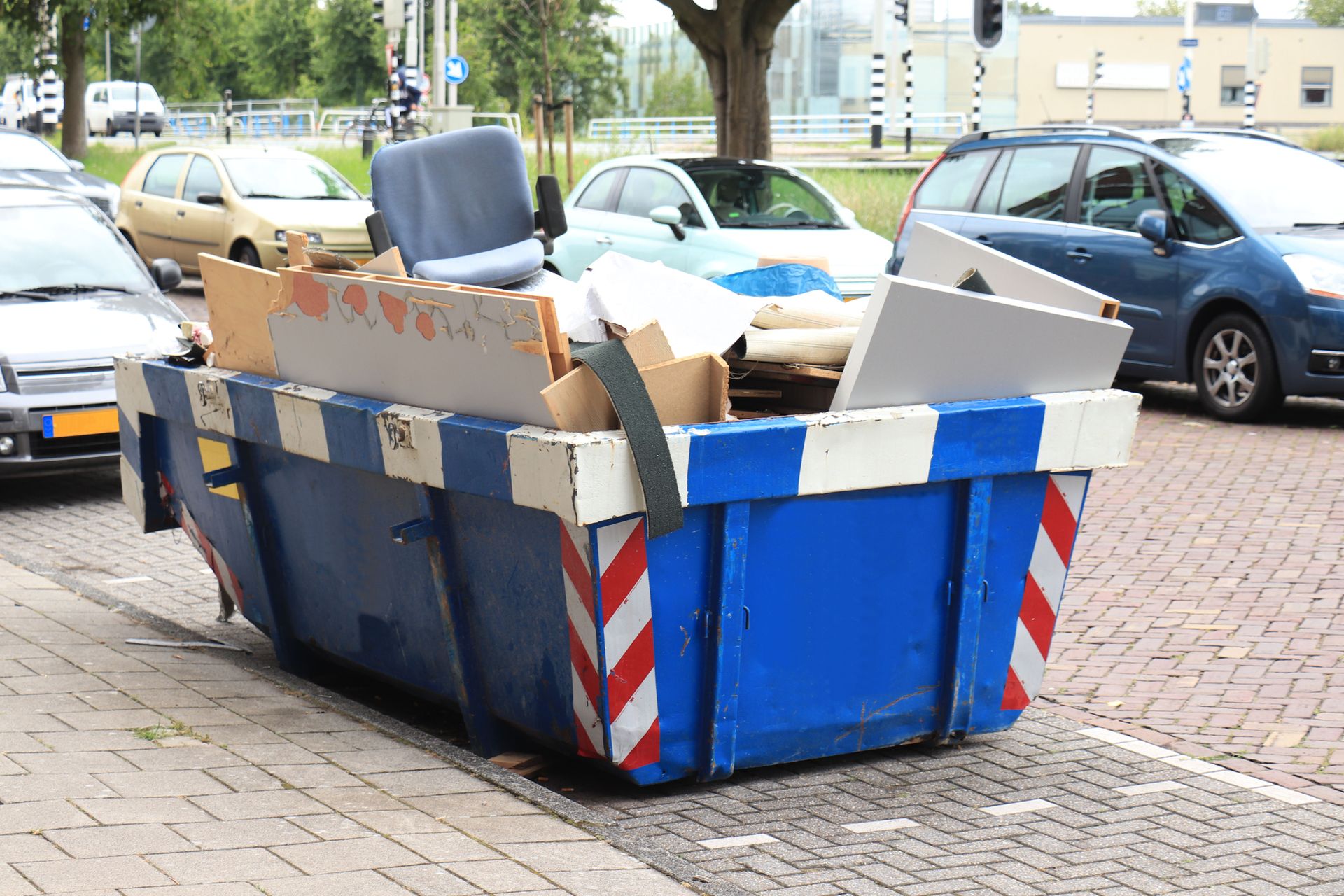 Blue dumpster filled with cardboard, furniture, and trash on a sidewalk beside parked cars