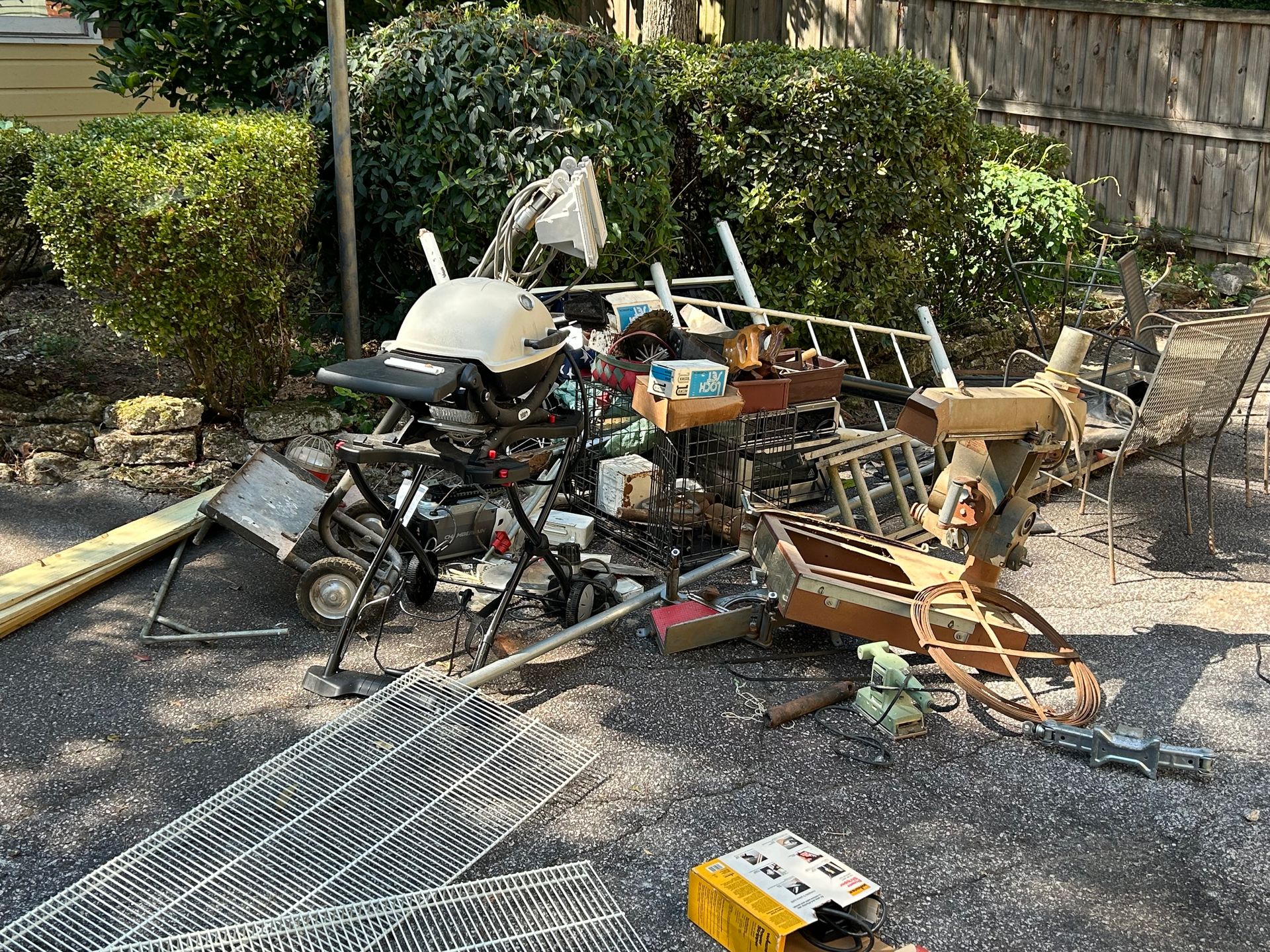 Piles of damaged debris and metal parts scattered on gravel beside a hedge in bright sunlight