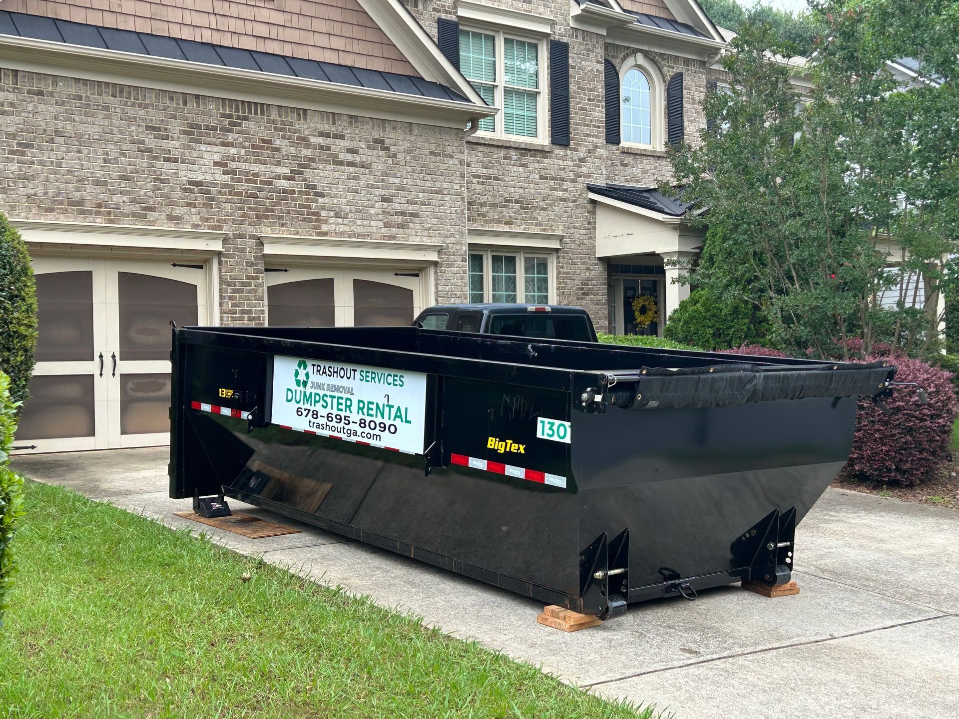 Blue dumpster filled with cardboard, furniture, and trash on a sidewalk beside parked cars
