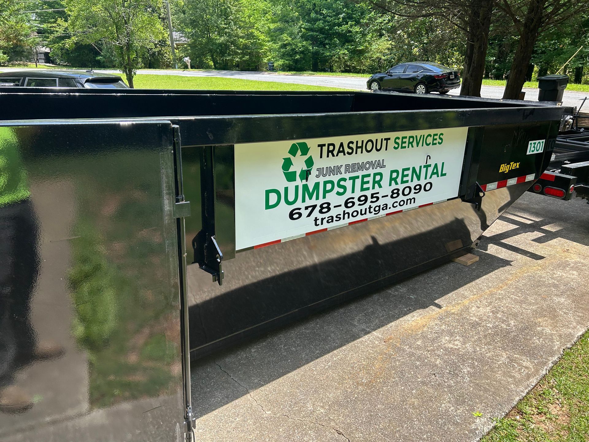 Black dumpster with Trashit Services dumpster rental sign on a sidewalk near trees