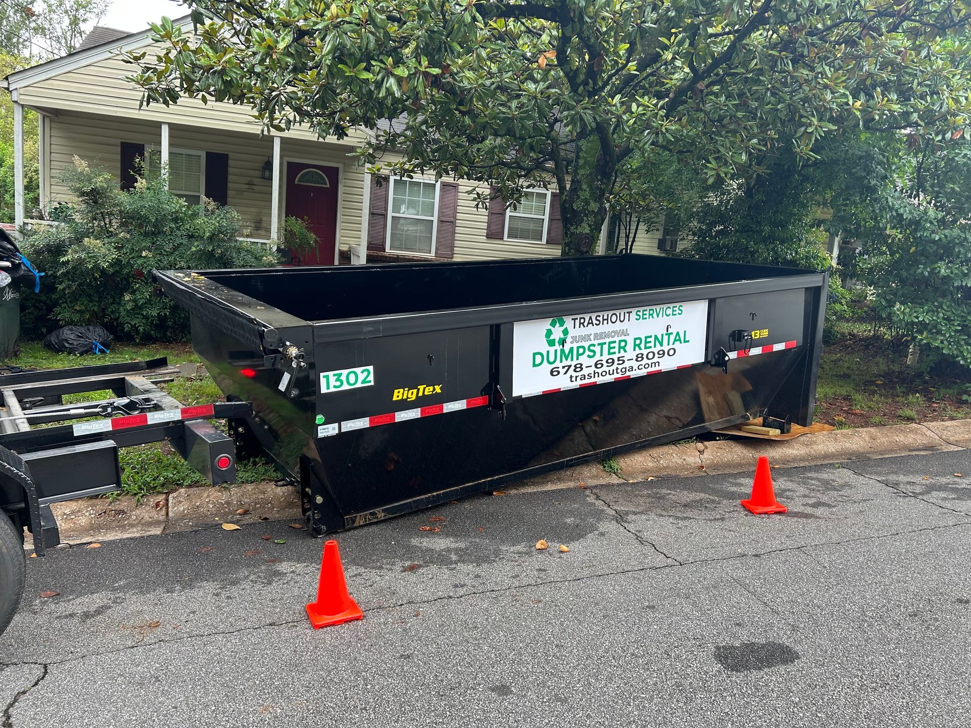 Black dumpster in front of a house with orange cones on a residential street