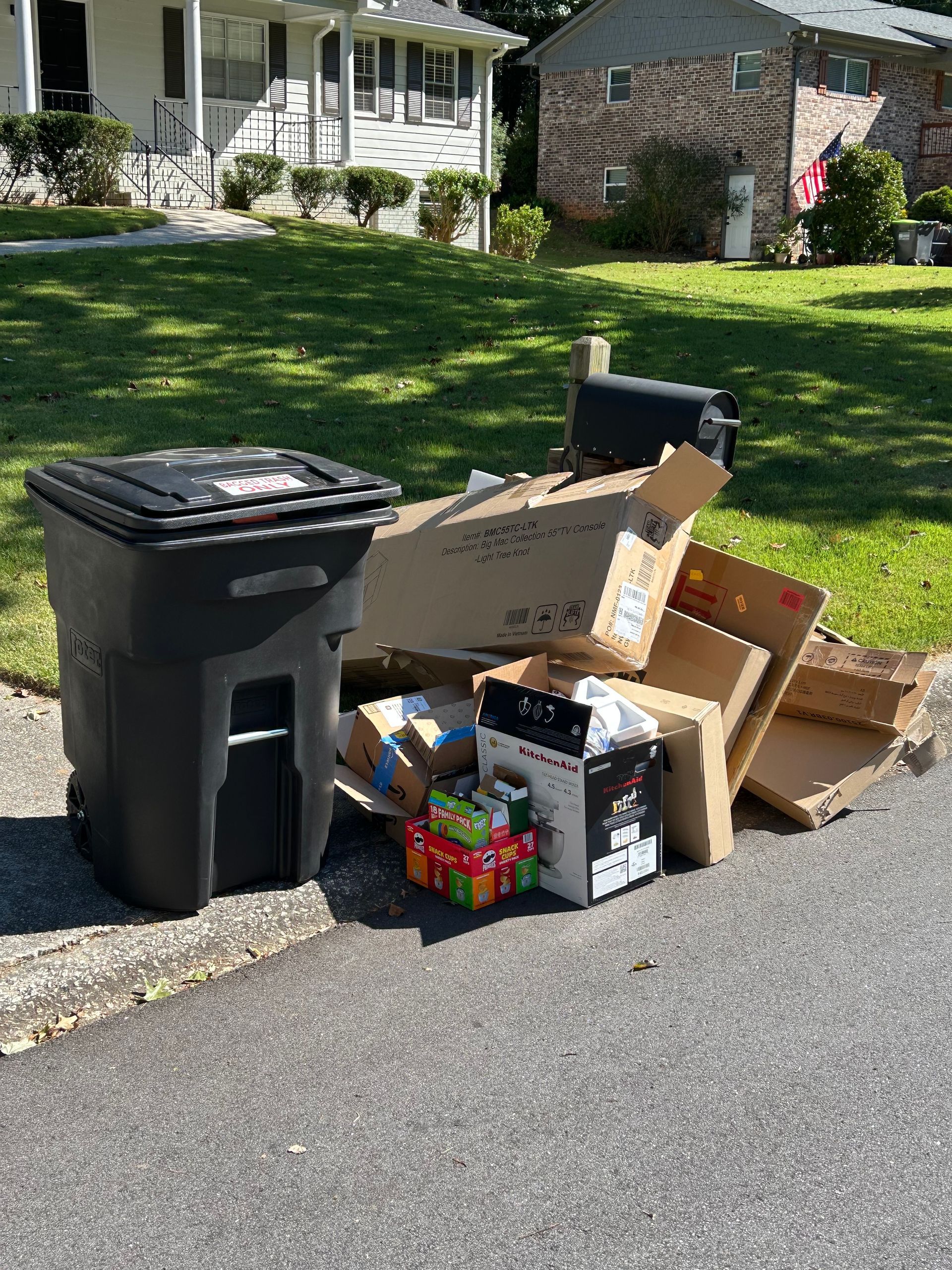 Garbage bin beside curb with cardboard, recycling, and boxes piled for pickup on a suburban street