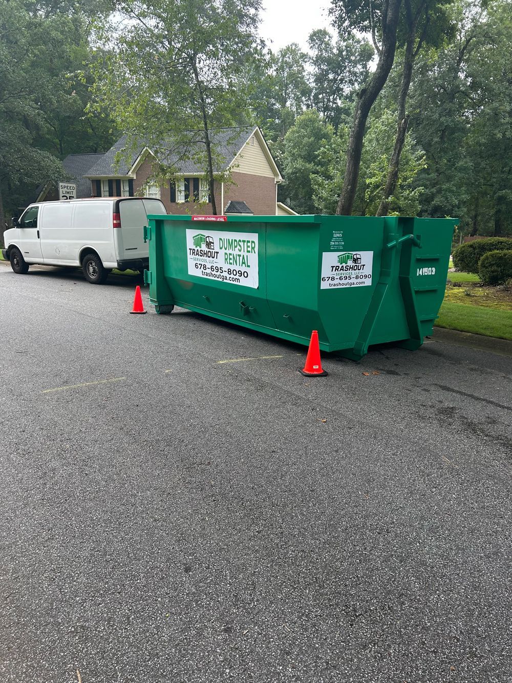 Blue dumpster filled with cardboard, furniture, and trash on a sidewalk beside parked cars