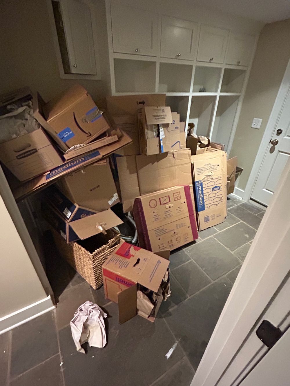 Cluttered hallway with stacked moving boxes and bags near a white door and cabinets