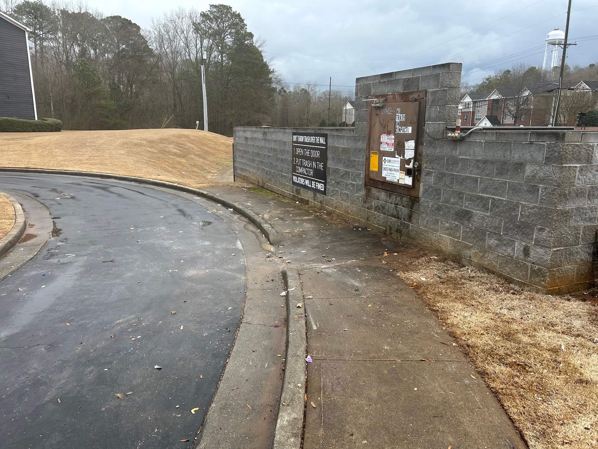 Wet curved road beside stone wall and gate, with signs and muddy gravel area on a cloudy day