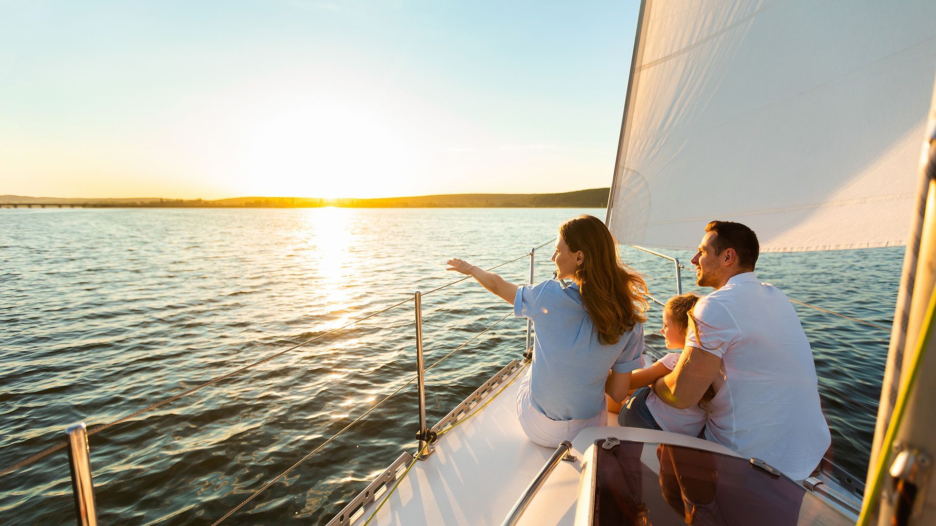 A family sits on the deck of a sailboat, watching the sunset over the water as one person points toward the horizon.
