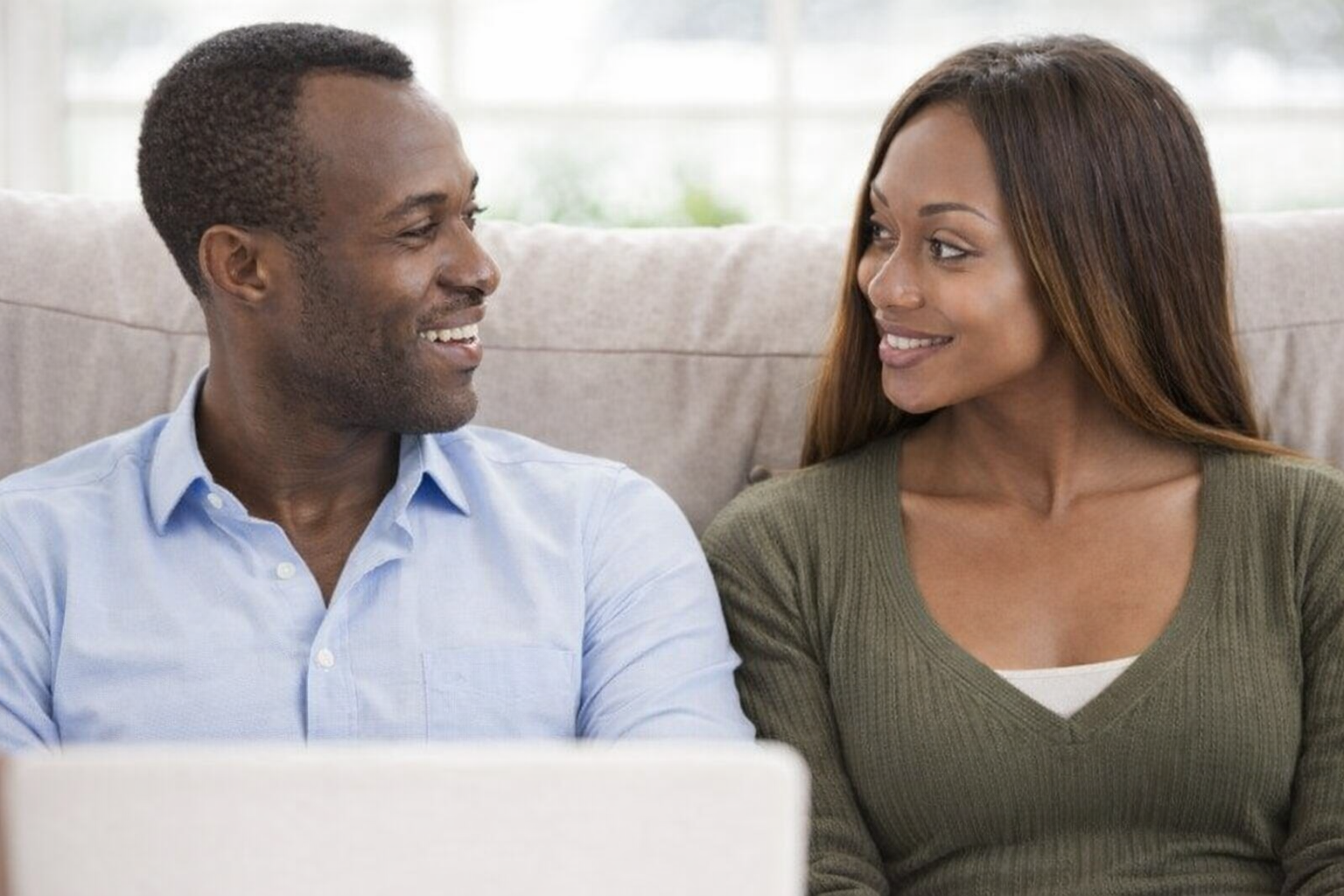 Two people sit side-by-side on a sofa, looking at each other and smiling while using a laptop.