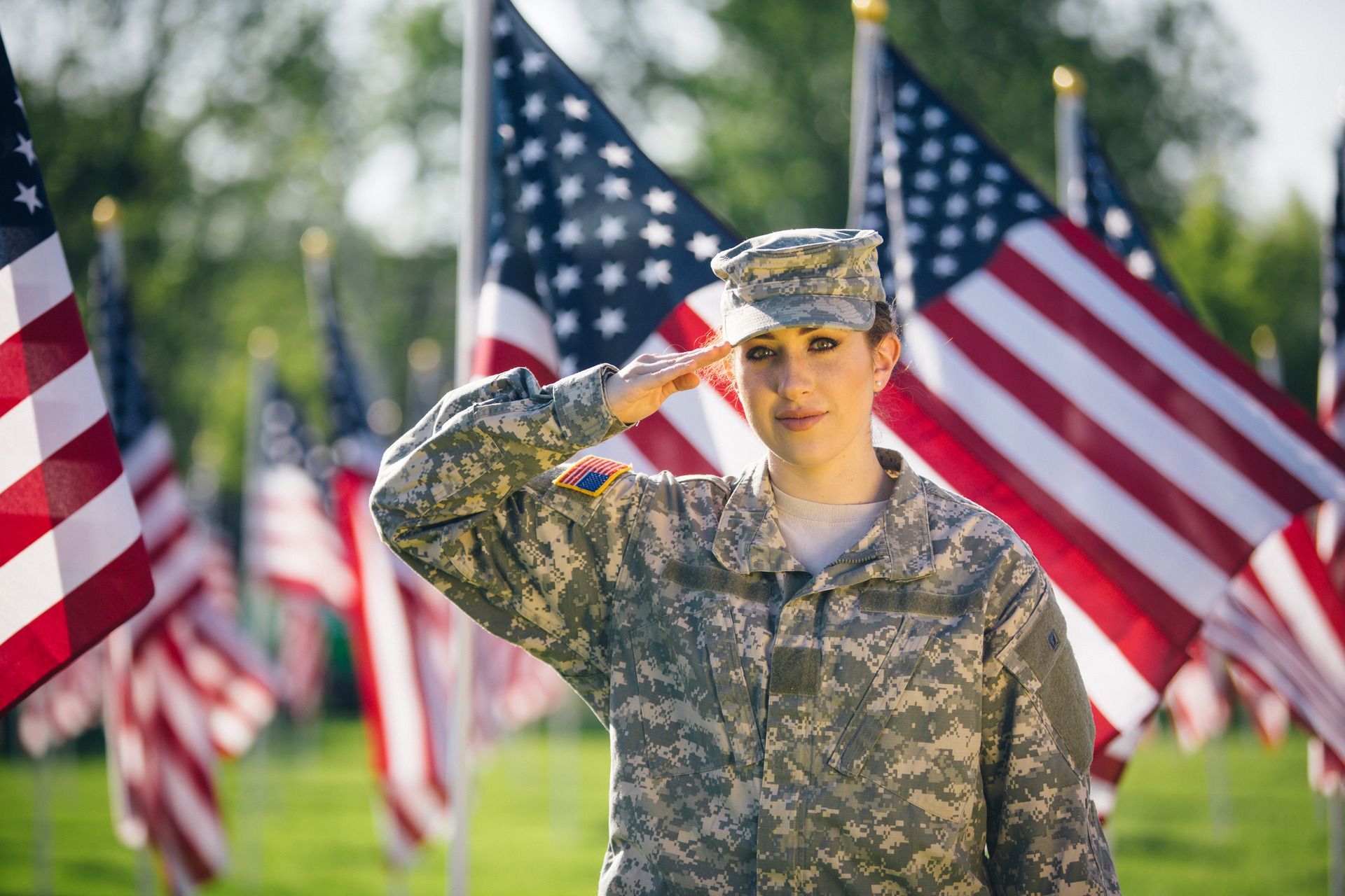 A service member in camouflage uniform salutes, positioned in a field filled with many American flags.