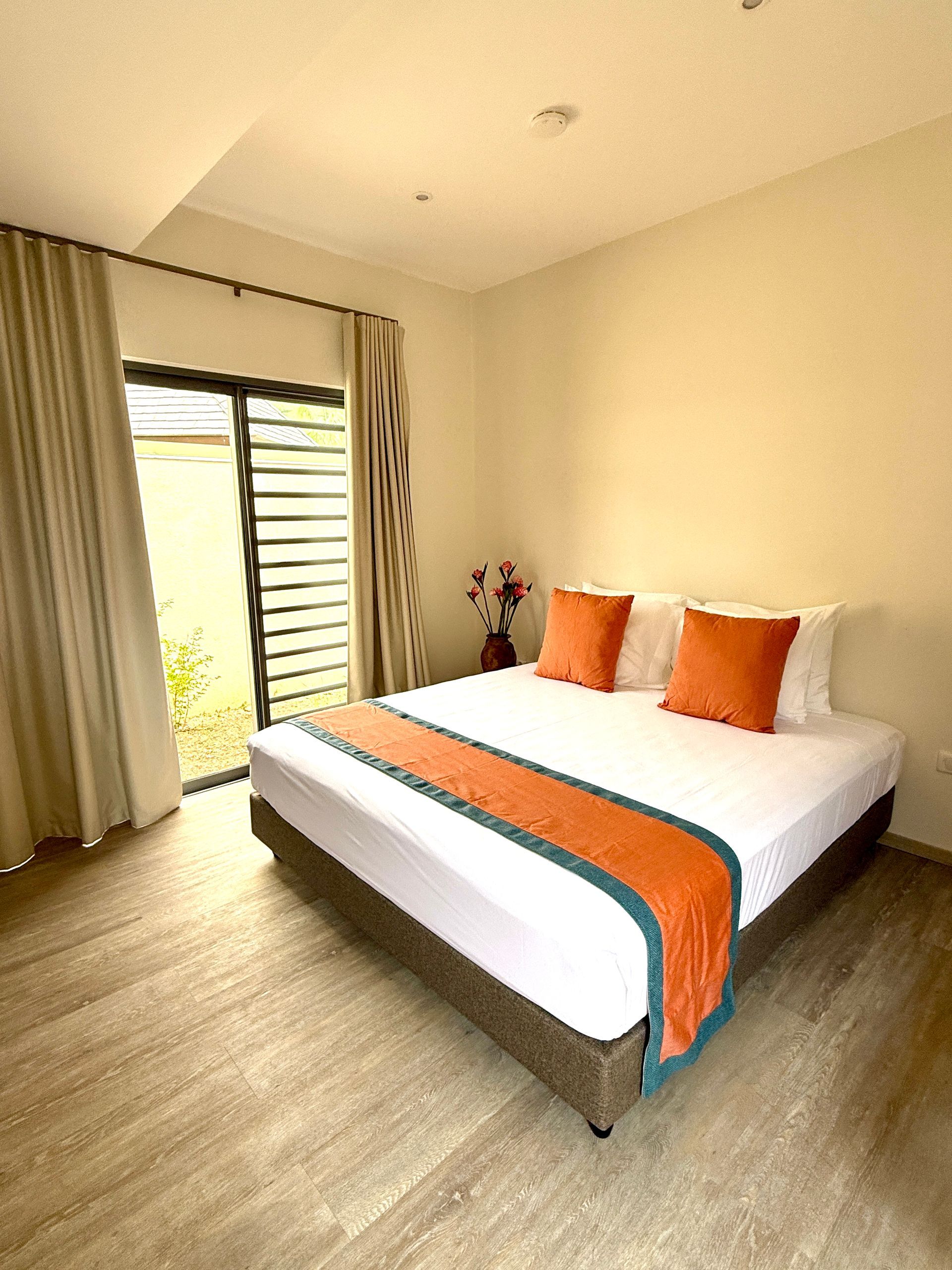 Bedroom with a bed, orange pillows and runner, neutral walls, and a sliding glass door.