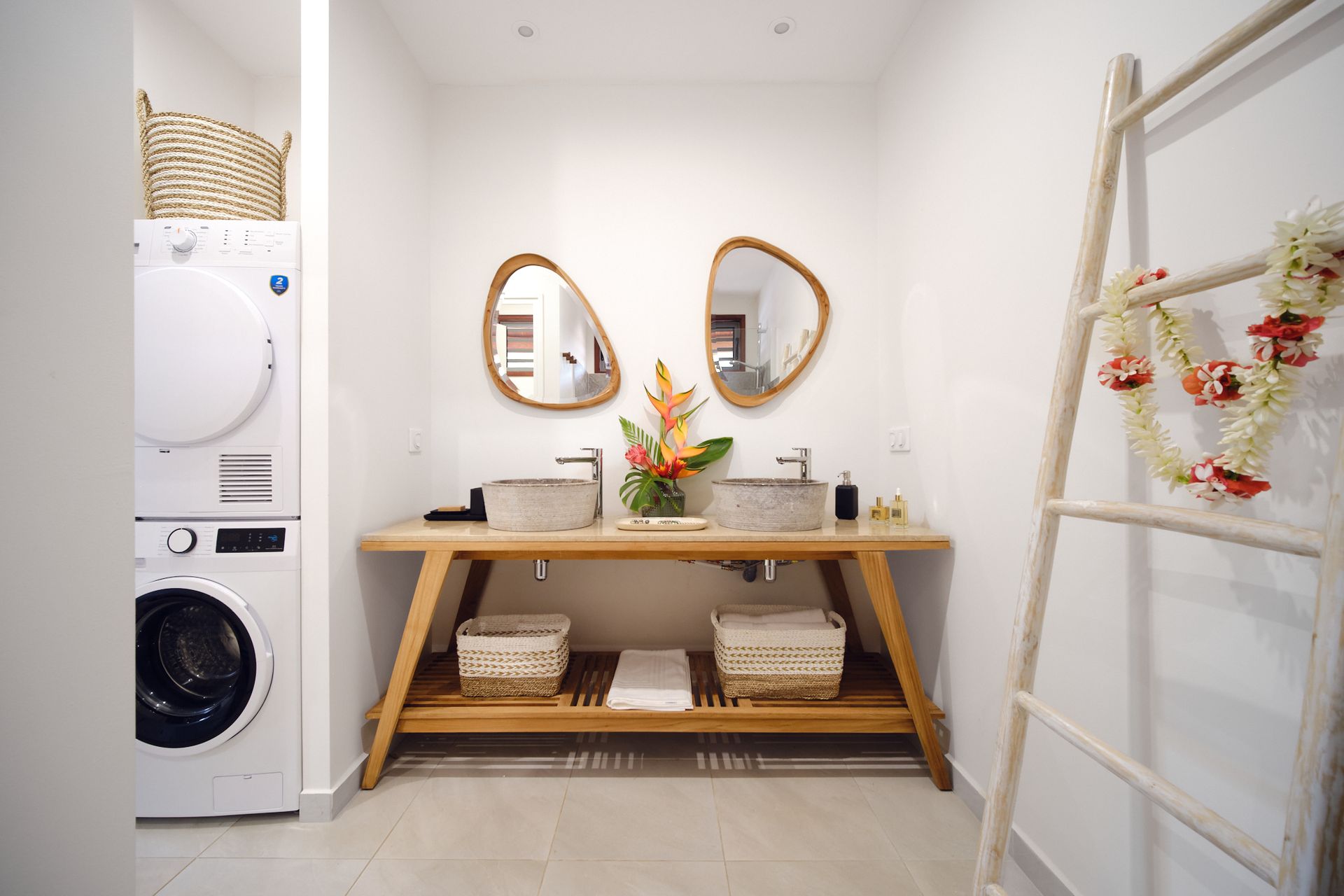 Bathroom with double sinks, wooden vanity, mirrors, and stacked washer/dryer; decor includes a ladder and baskets.
