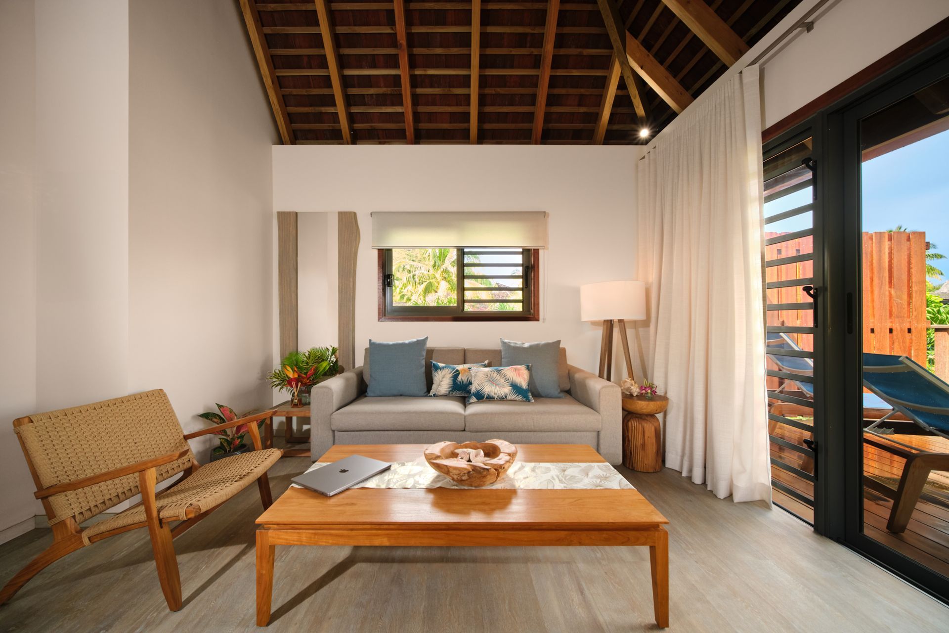 Living room with couch, coffee table, woven chair, wood-beamed ceiling, and balcony access.