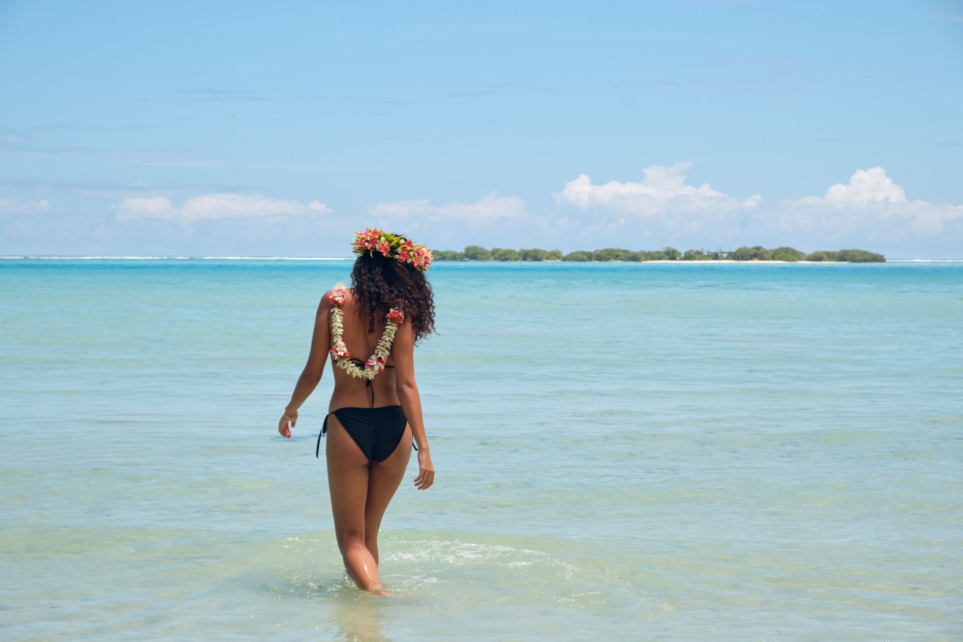 Woman in black bikini and flower leis walks into calm, turquoise ocean. Small island visible in distance.