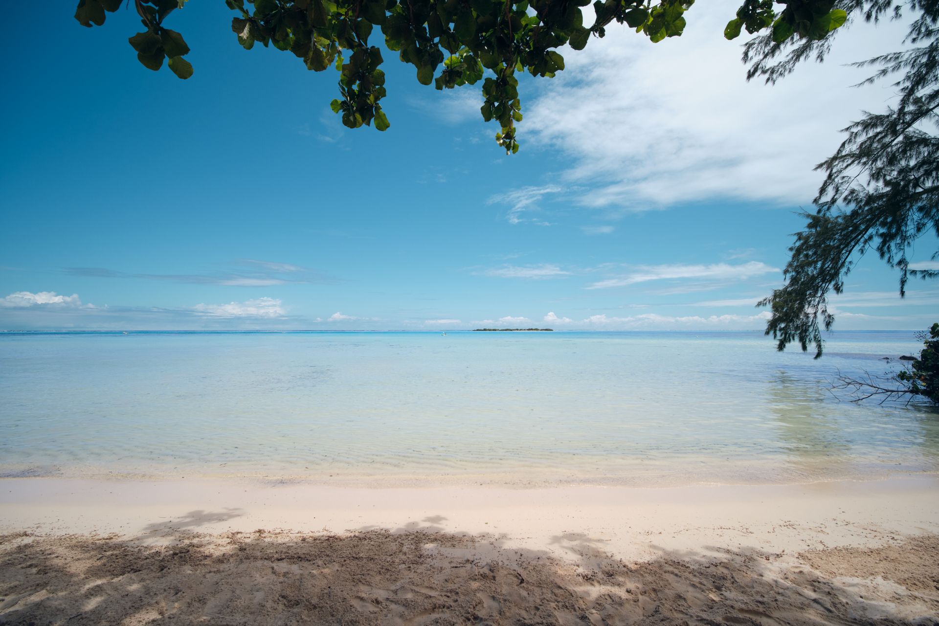 Beach scene with light sand, clear water, blue sky, and a small island in the distance.
