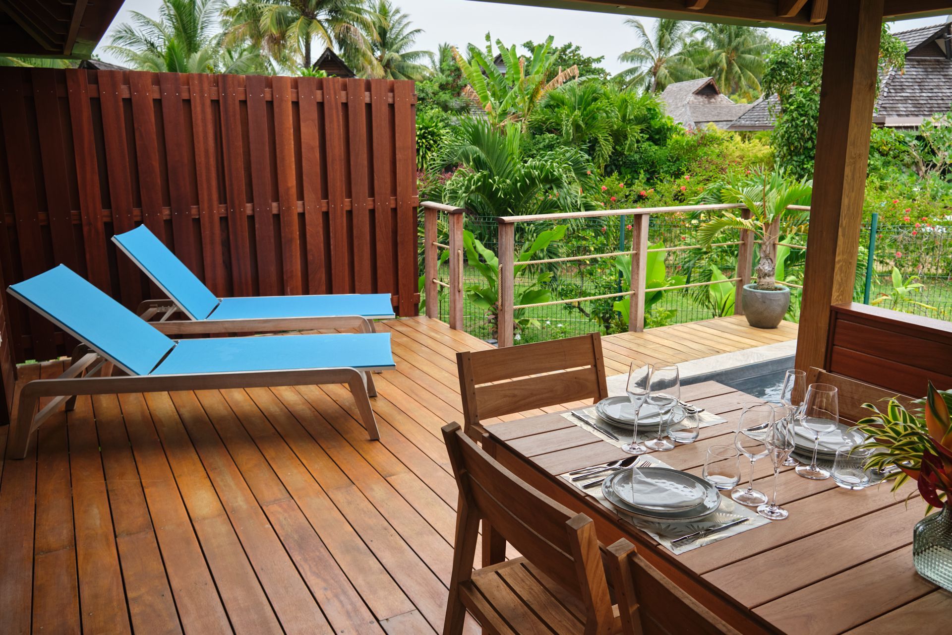 Wooden deck with two blue lounge chairs, table set for dining, and lush green tropical foliage.
